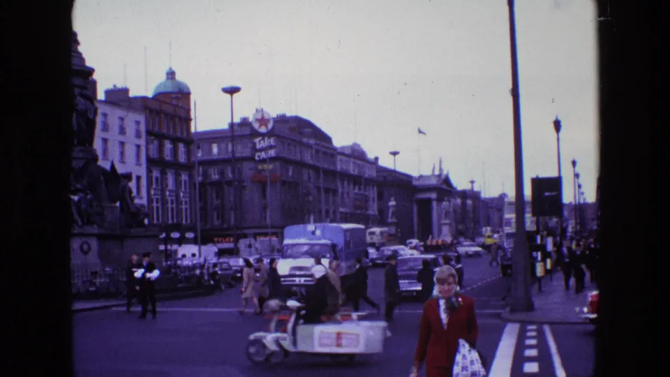 1969 Ireland home-movie view of pedestrians crossing among cars and vans near a large city monument and commercial signage.