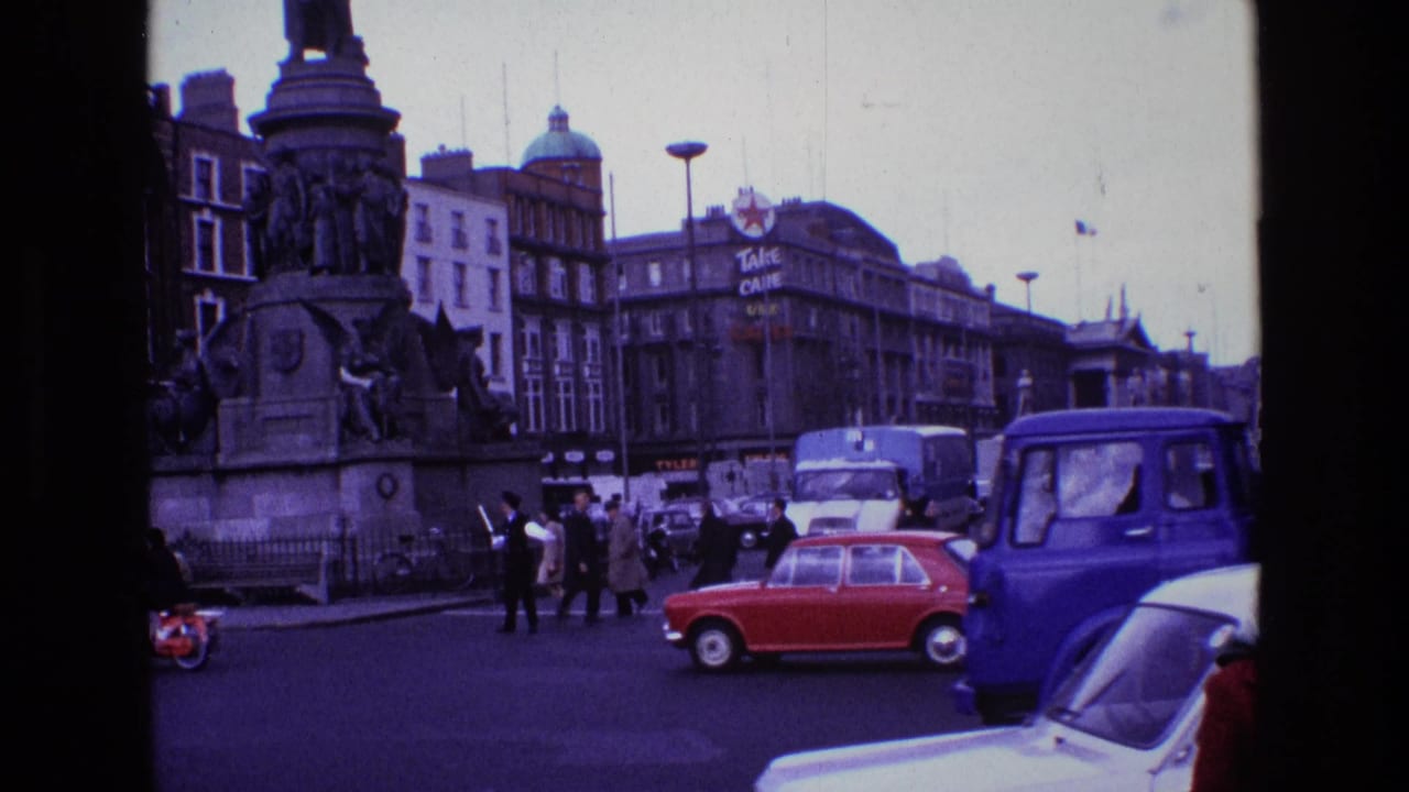 Vehicles move through a 1969 Ireland city intersection, with a monument, pedestrians, and background commercial signage visible.