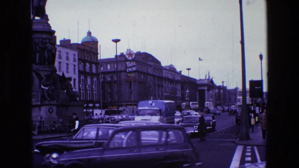 A classic dark car passes close to the camera in a 1969 Ireland street scene with traffic and pedestrians beyond.