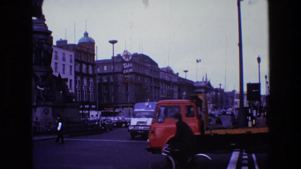 A red truck crosses a 1969 Ireland city intersection in front of cars, pedestrians, street lamps, and a large monument.