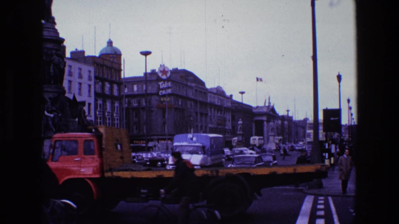 1969 8mm street view in Ireland showing classic cars, a red flatbed truck, pedestrians, buses, and large building signs under an overcast sky.