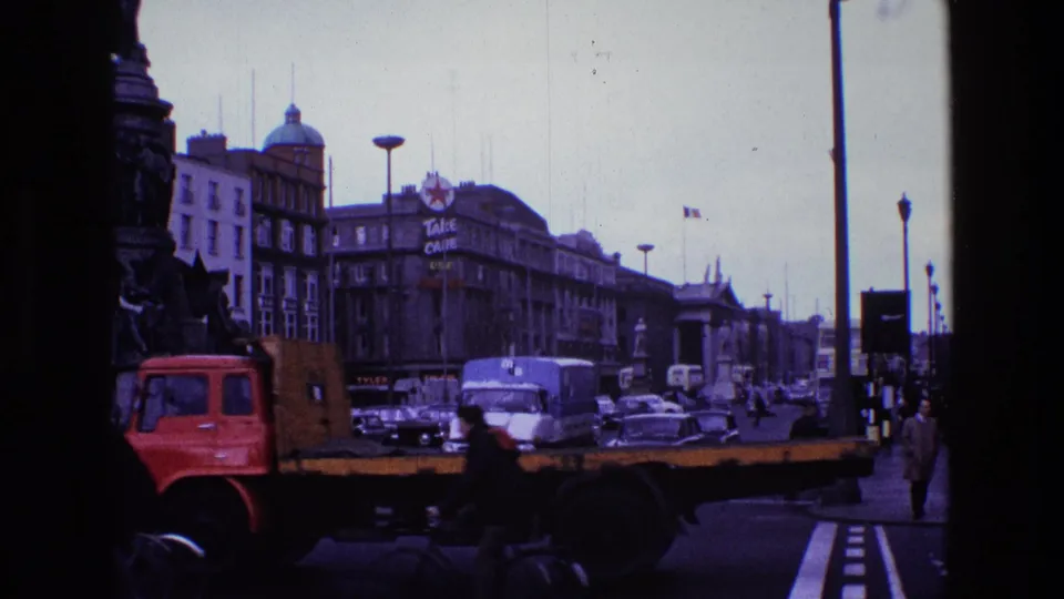 1969 8mm street view in Ireland showing classic cars, a red flatbed truck, pedestrians, buses, and large building signs under an overcast sky.