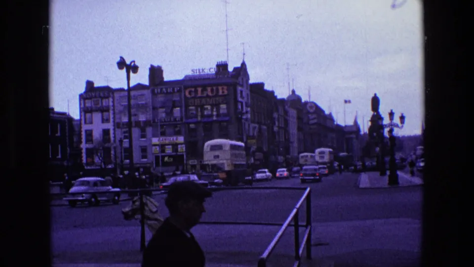 1969 Ireland street intersection with buses, classic cars, a pedestrian in silhouette, railings, ornate lamps, and prominent commercial signage.