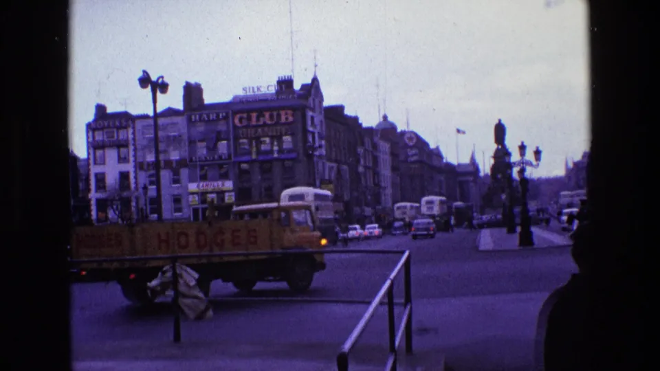 1969 Ireland traffic scene with a yellow flatbed truck moving past buses, cars, pedestrians, railings, and large commercial signs.