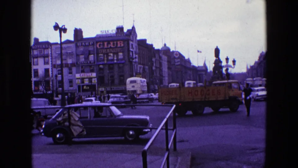 1969 Ireland street view showing a black car, yellow flatbed truck, buses, pedestrians, railings, ornate lamps, and commercial signage.