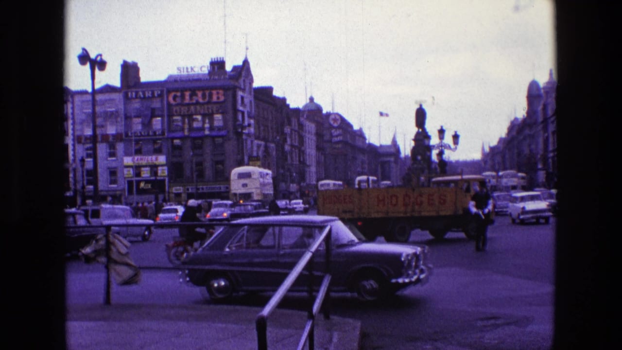 1969 Ireland street scene with classic cars, buses, pedestrians, shop signs, a yellow delivery truck, and a statue in a busy intersection.