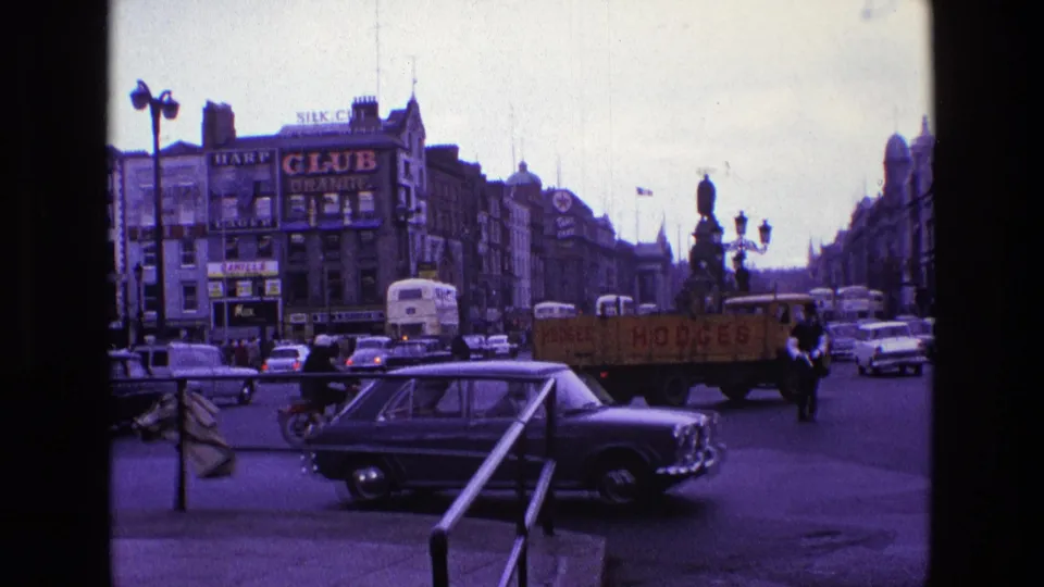 1969 Ireland street scene with classic cars, buses, pedestrians, shop signs, a yellow delivery truck, and a statue in a busy intersection.