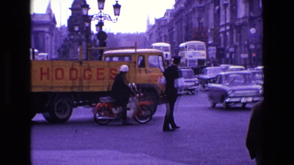 1969 Ireland traffic scene with a uniformed officer, motorcyclist, bicycle, yellow HODGES truck, buses, and classic cars in view.