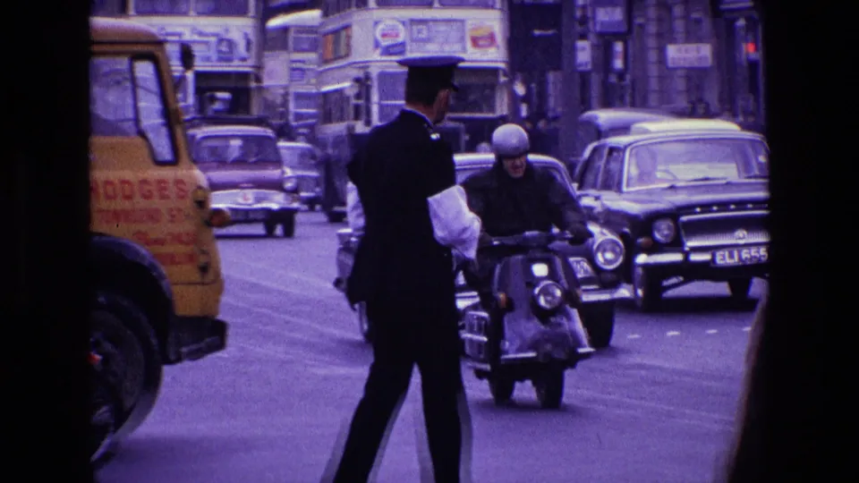 1969 Ireland street traffic with a uniformed officer, motorcyclist, classic cars, double-decker bus, yellow truck edge, and visible car plate.