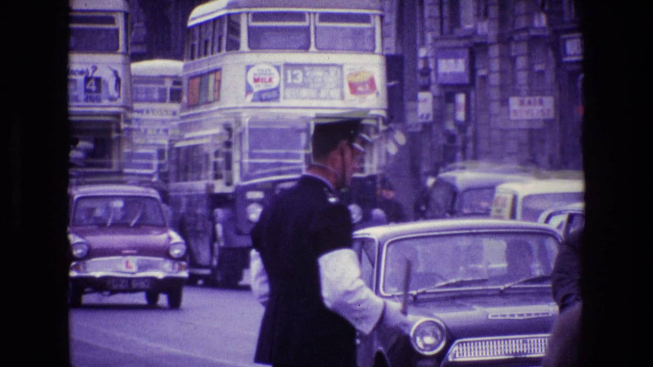 Policeman in uniform directs traffic beside vintage cars and double-decker buses.