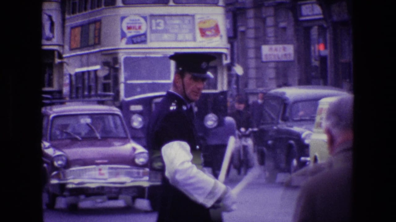 Traffic policeman faces across the road with buses, cars, and pedestrians behind him.