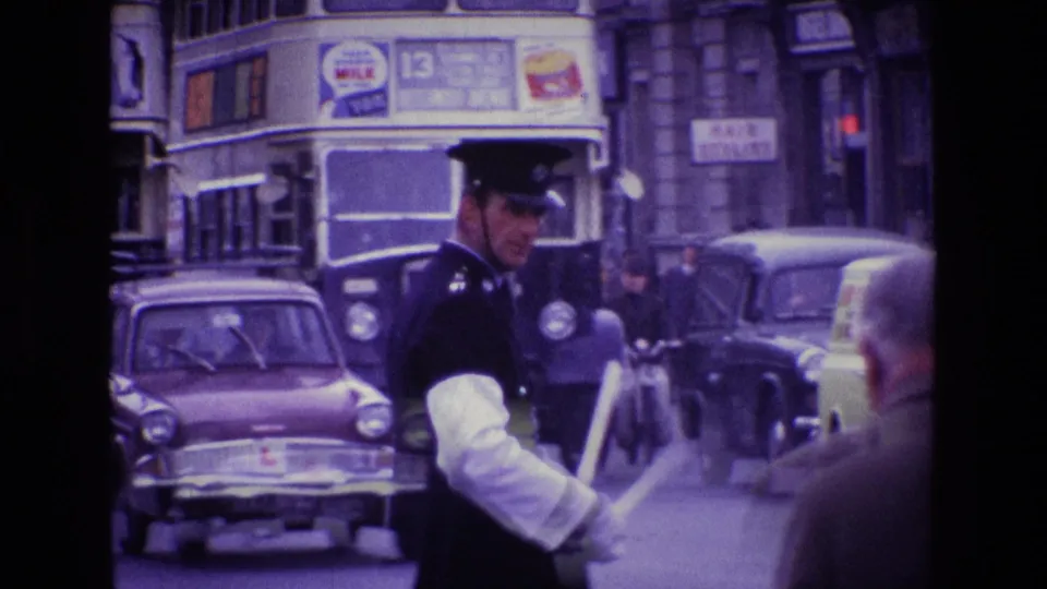 Uniformed policeman in a 1969 Irish city traffic scene, with buses, cars, pedestrians, and shopfronts or building facades behind him.