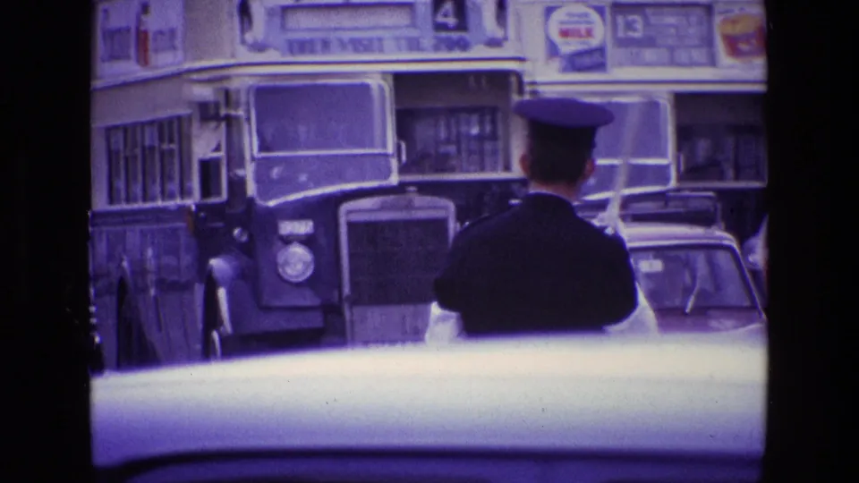 Rear view of a traffic policeman in Ireland, 1969, standing before oncoming double-decker buses and mid-century cars.