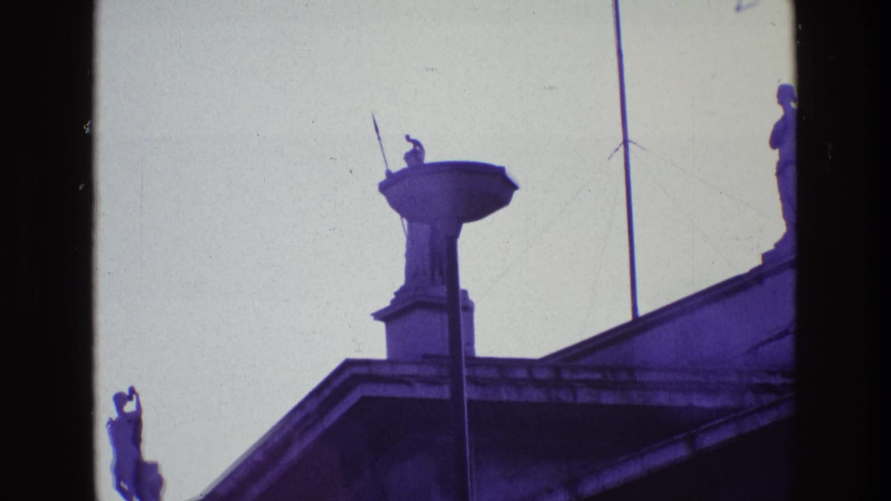 1969 Ireland 8mm frame showing a spear-bearing guardian statue on a rooftop pedestal, seen from below against a pale sky.