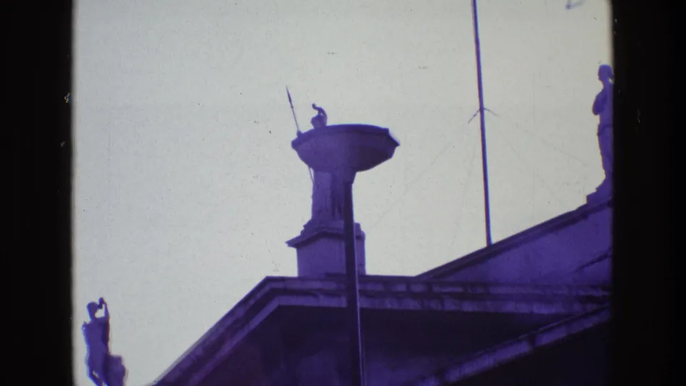 1969 Ireland 8mm frame showing a spear-bearing guardian statue on a rooftop pedestal, seen from below against a pale sky.