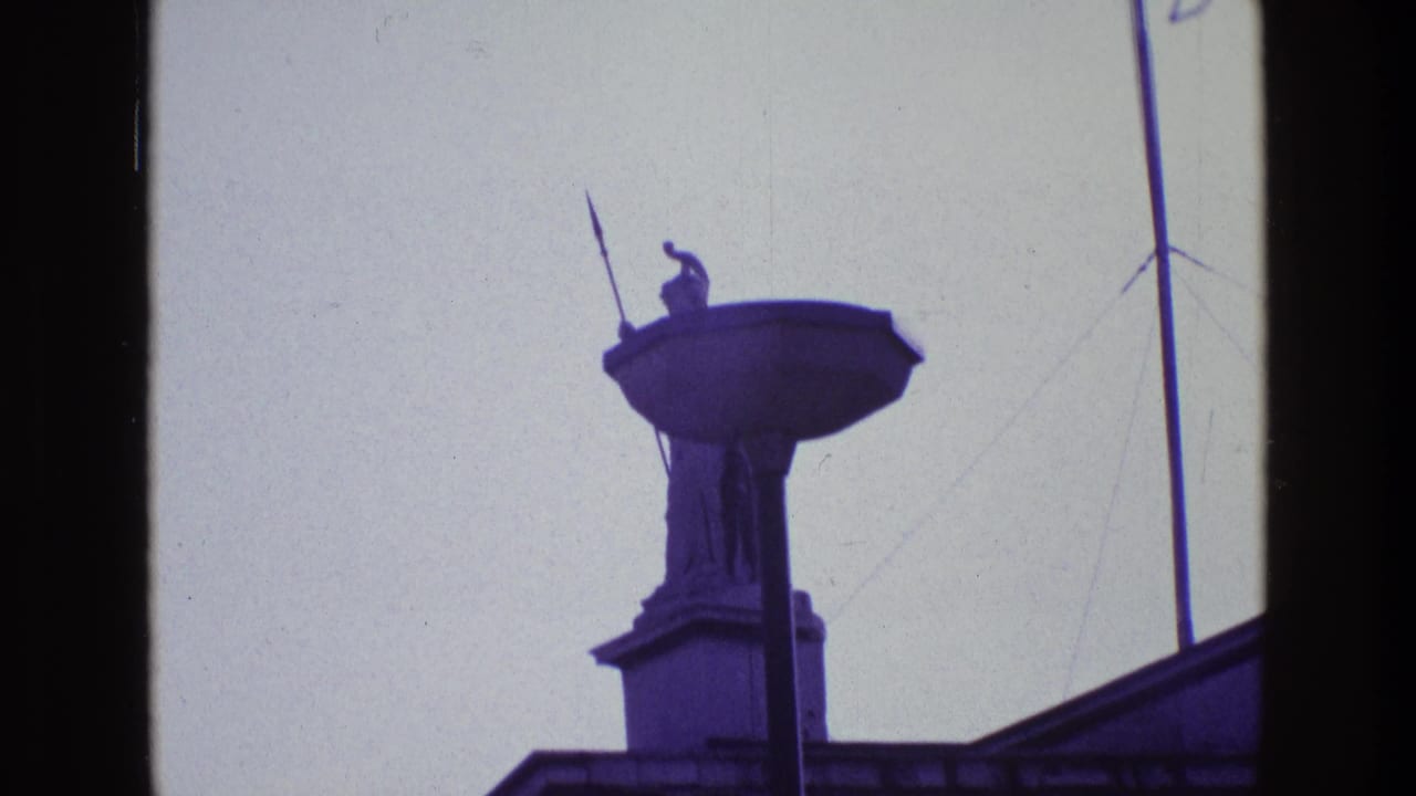 1969 Ireland home-movie frame with the guardian statue and spear more centered, rising above a dark roof edge.