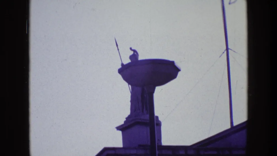 1969 Ireland home-movie frame with the guardian statue and spear more centered, rising above a dark roof edge.