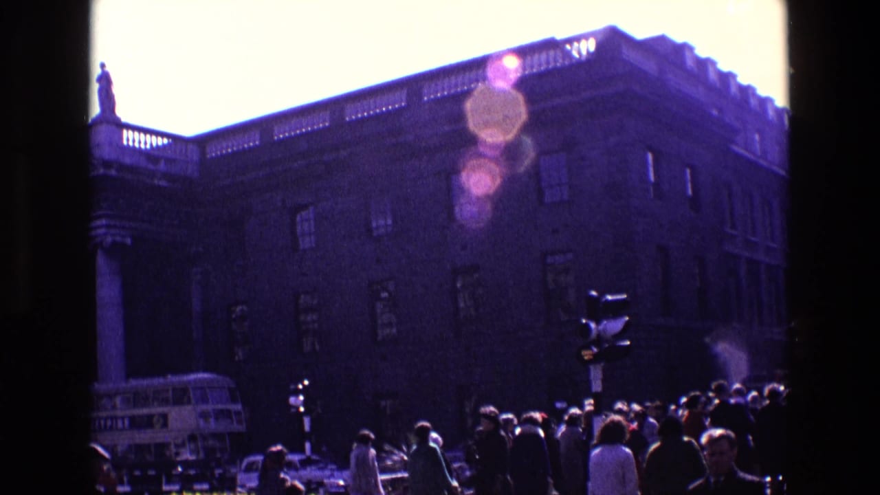 1969 Ireland street view with a large columned building, rooftop statue, crowd of pedestrians, traffic signal, and double-decker bus.