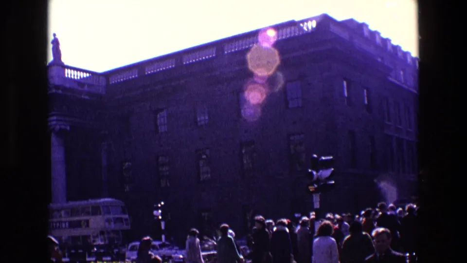 1969 Ireland street view with a large columned building, rooftop statue, crowd of pedestrians, traffic signal, and double-decker bus.