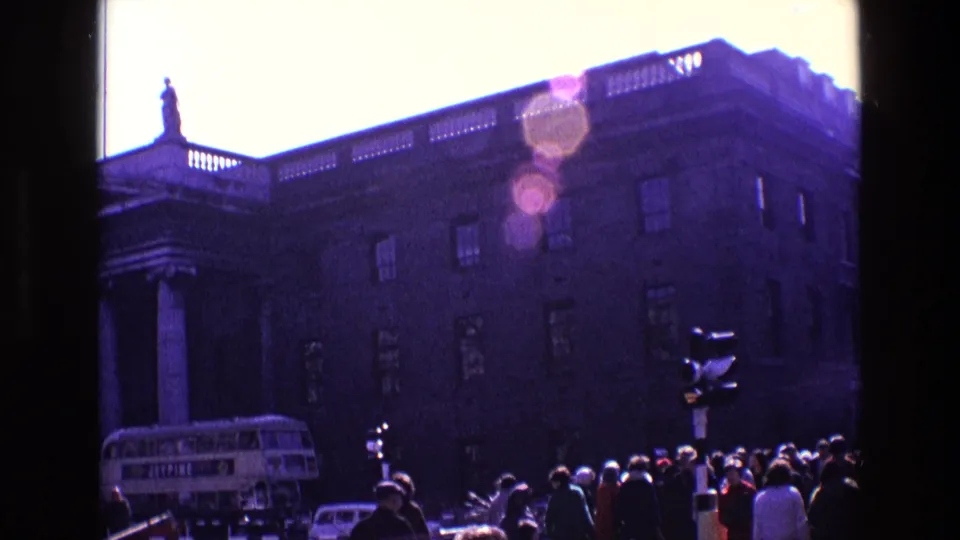 1969 Ireland 8mm frame of a crowded city street, large formal building, columns, rooftop statue, bus, cars, and bright lens flare.