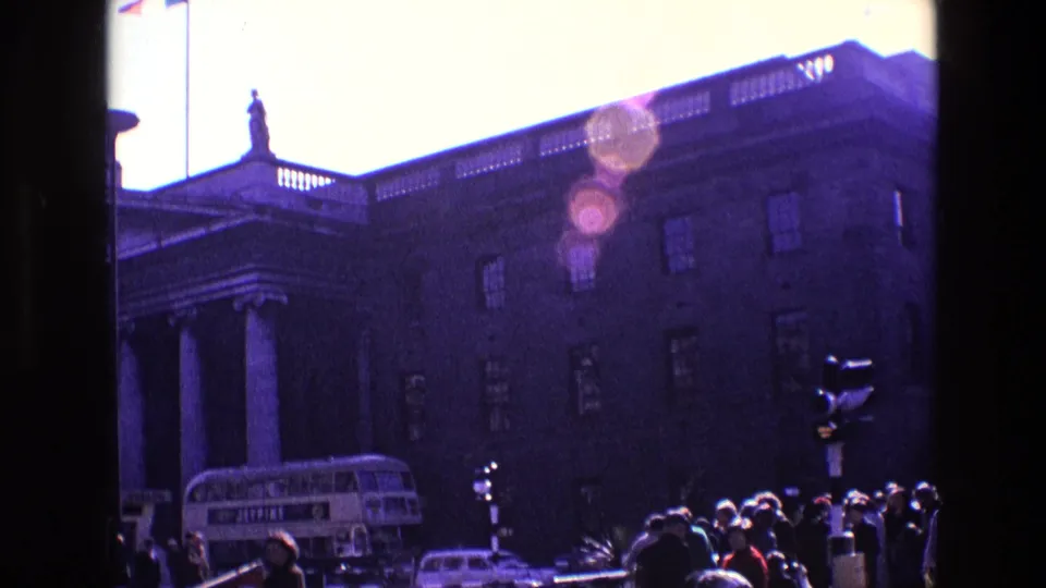 1969 Ireland street scene with pedestrians, traffic signal, double-decker bus, cars, and a large columned building under strong sun flare.