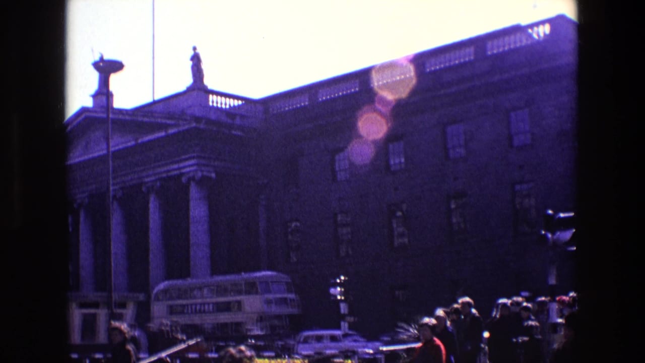 1969 Ireland home-movie frame showing a columned public building, rooftop statues, pedestrians and a double-decker bus on a busy street.
