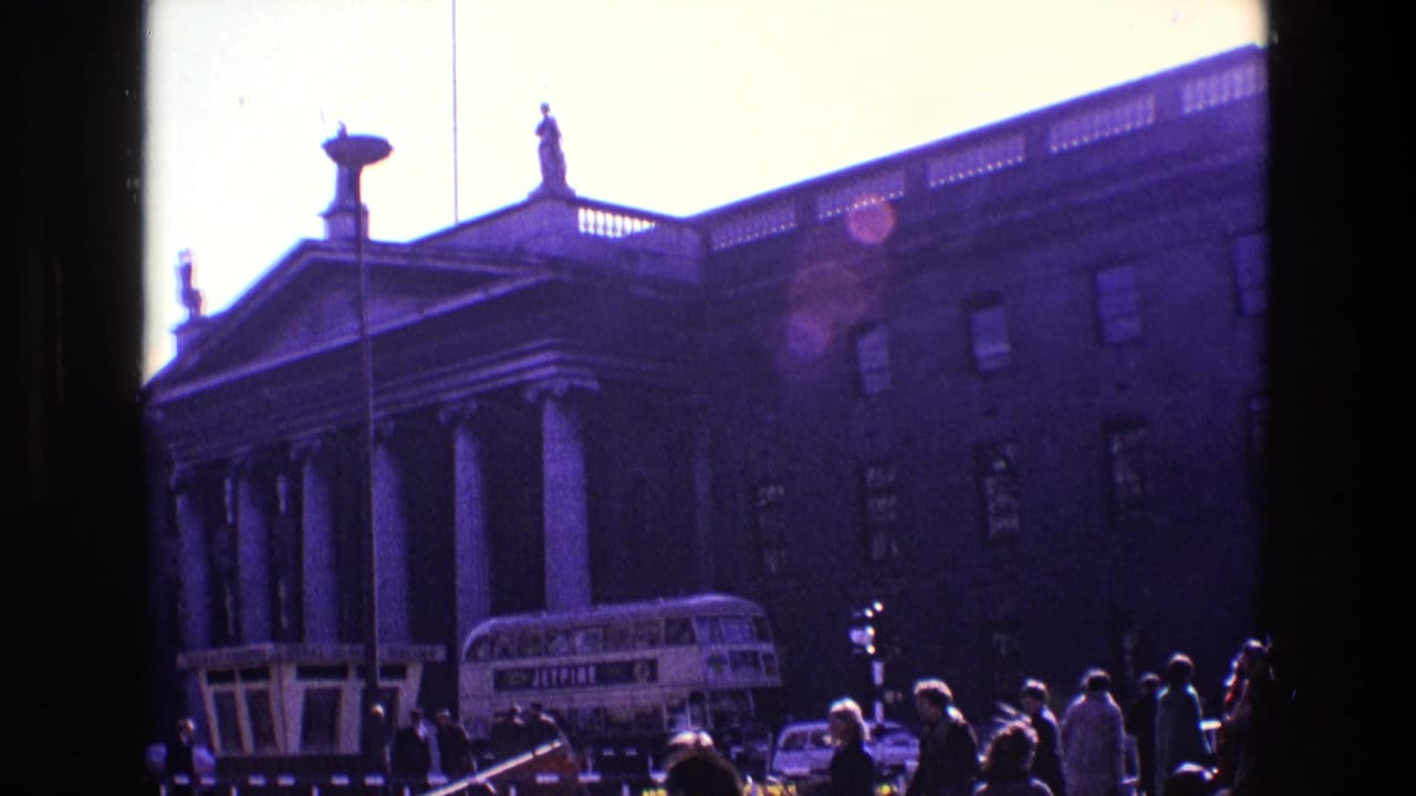 Pedestrians and traffic near a columned building with a double-decker bus at the curb.