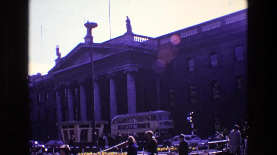 1969 Ireland home-movie angle on a classical building with columns, a double-decker bus, cars and pedestrians under bright backlight.