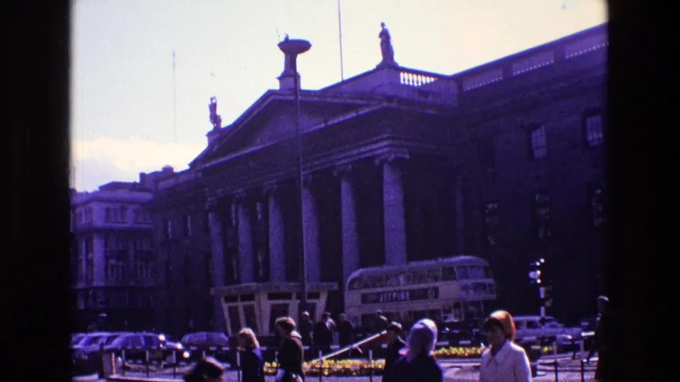 1969 Ireland frame showing a classical street frontage with statues, a double-decker bus, cars, pedestrians and a low flower bed.