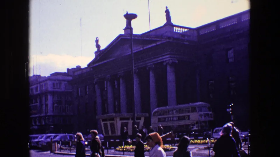 1969 Ireland street view with pedestrians, vintage cars, a bus, and a large neoclassical building with columns and roof statues.