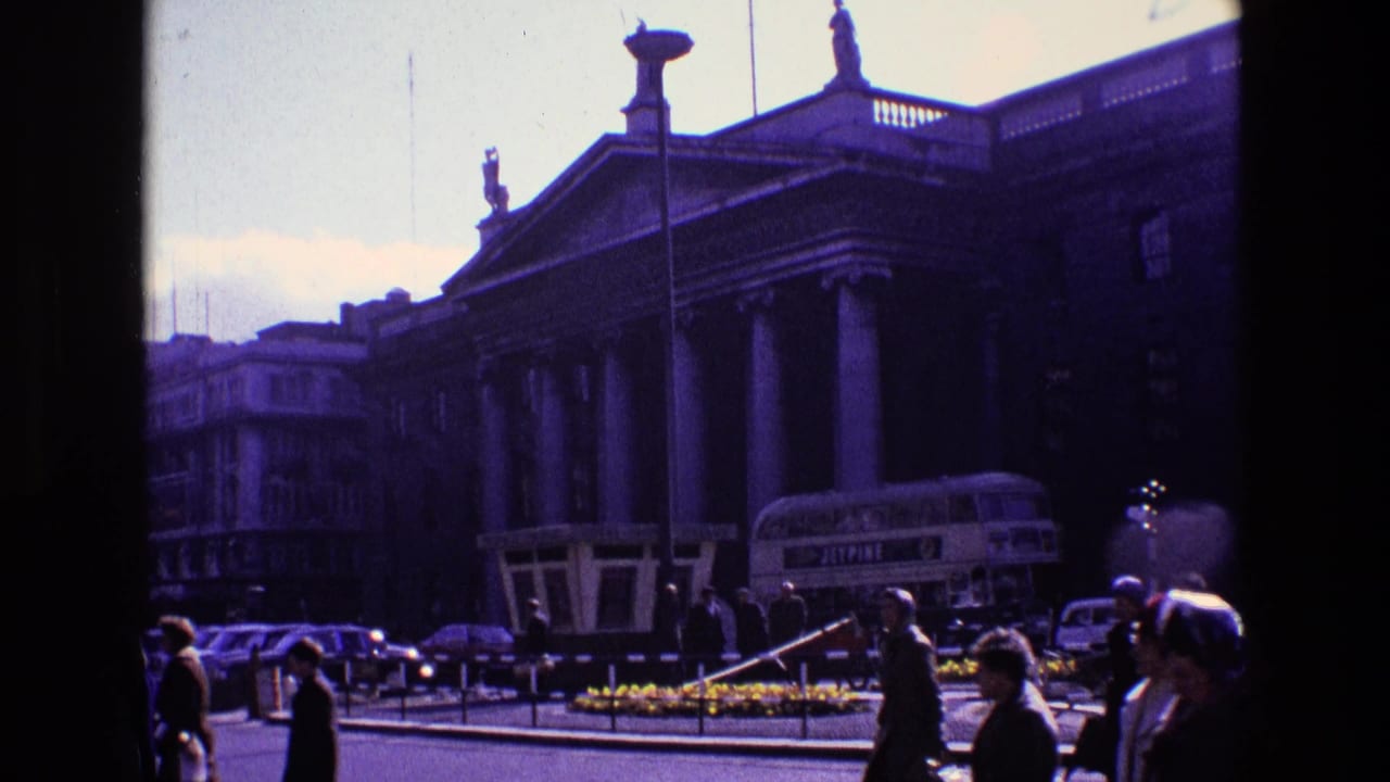 Crowds walk along the street as a bus and cars pass a large columned building.