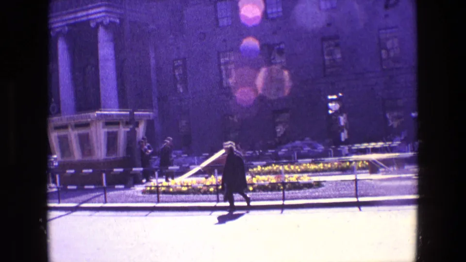 A sun-flared 1969 Ireland street frame shows a person walking by flower beds, railings, cars, and a large public building.