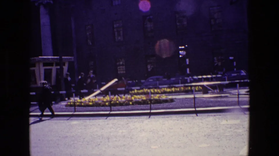 1969 Ireland street view with pedestrians near a flower-lined median, traffic signals, railings, and a dark building facade behind them.