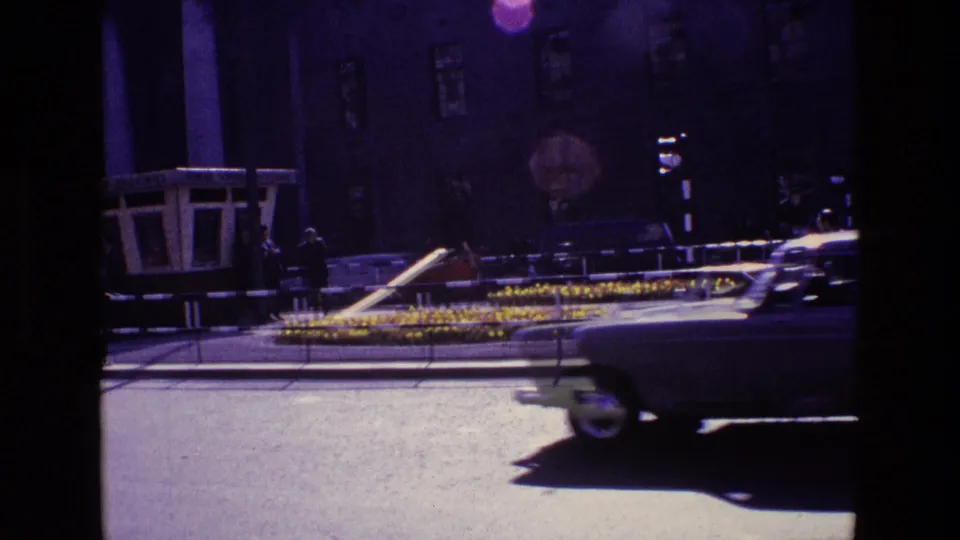 A light-colored classic car moves across a 1969 Ireland street view with a small kiosk, pedestrians, flower beds, railings, and traffic signals.