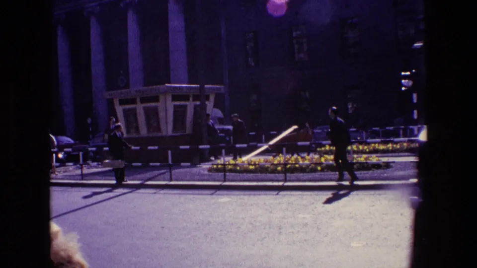 In the 1969 Ireland street scene, a man jogs past the flower-lined median while a kiosk, traffic lights, parked or passing cars, and a building sit behind him.