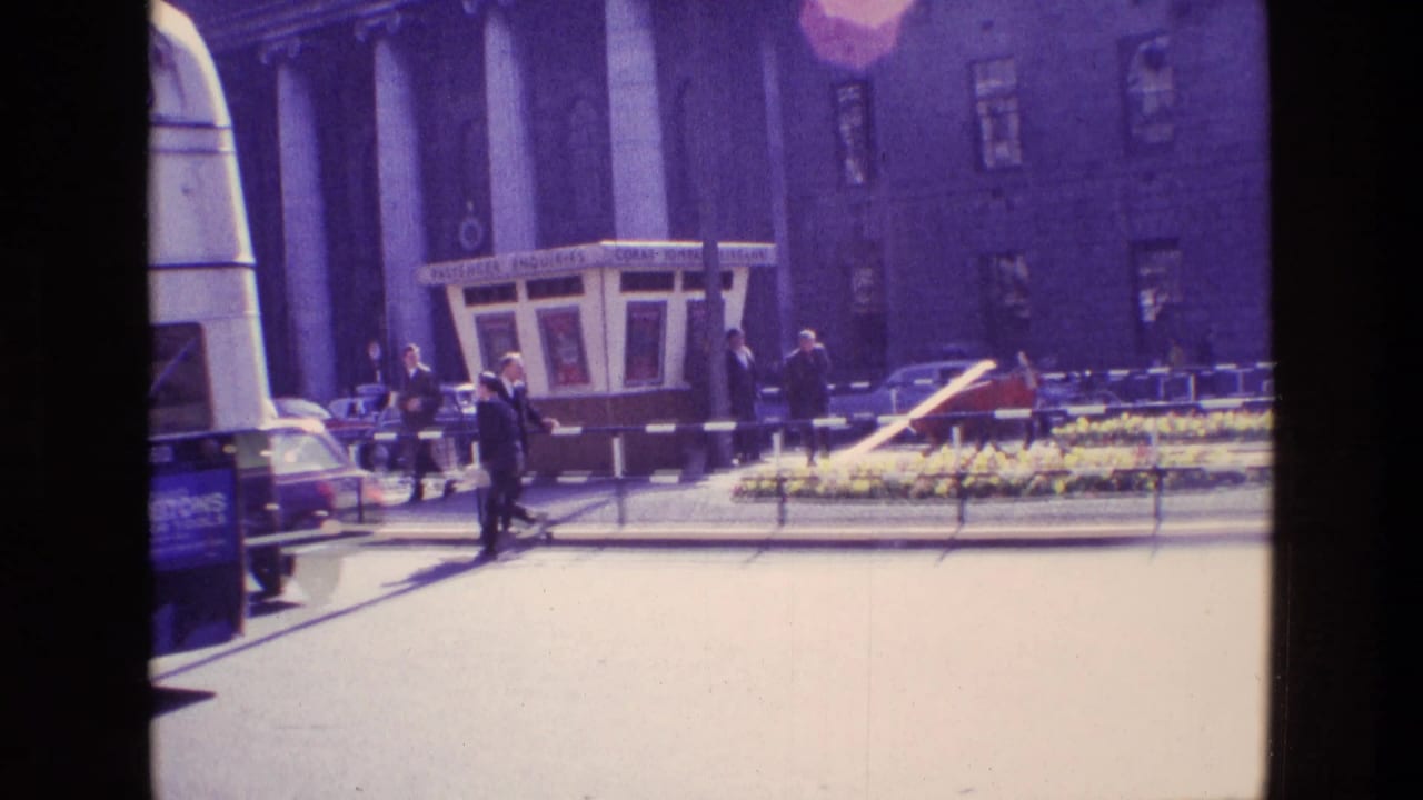 1969 Ireland street view with pedestrians, vintage cars, railings, and a flower bed outside a columned building.