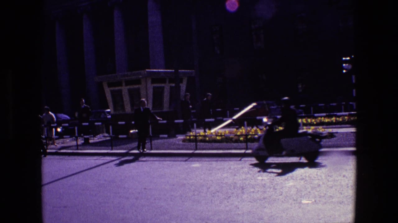 1969 Ireland city street with a scooter, pedestrians, railings, and a planted median near a formal building facade.