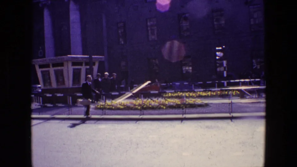 1969 Ireland street scene with a suited man crossing near a flower bed, railings, and a columned urban building.