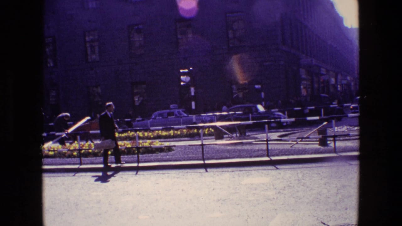 1969 Ireland street view with a suited pedestrian crossing near railings, a flower bed, older cars, and tall buildings in bright flare.