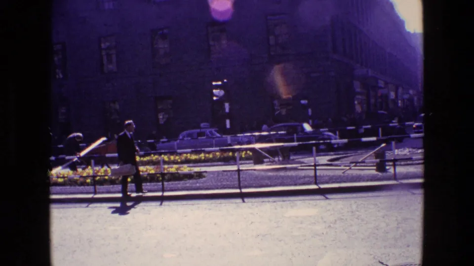 1969 Ireland street view with a suited pedestrian crossing near railings, a flower bed, older cars, and tall buildings in bright flare.