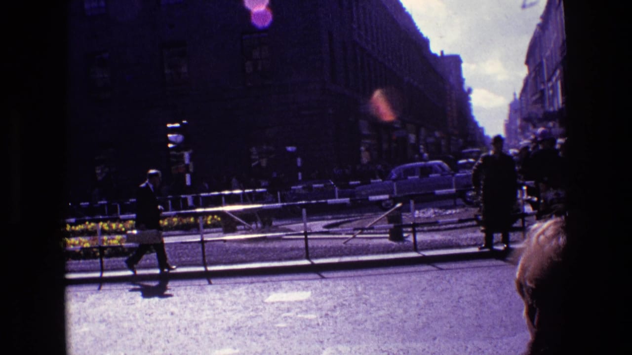 Pedestrians move along and across a busy street with older cars and railings behind them.