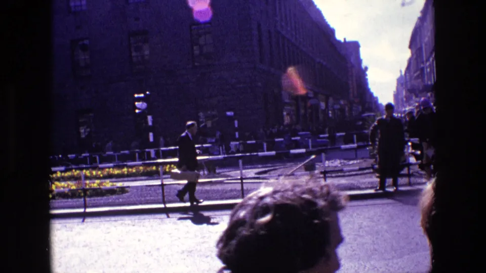1969 Ireland home-movie frame showing a suited pedestrian, curbside figures, street railings, flower bed, and urban buildings.