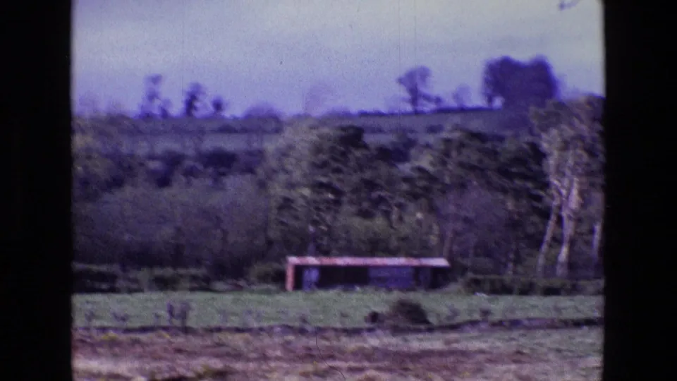 1969 Ireland 8mm frame showing a blurred rural field, trees, hedgerows, and a low red structure across the landscape.