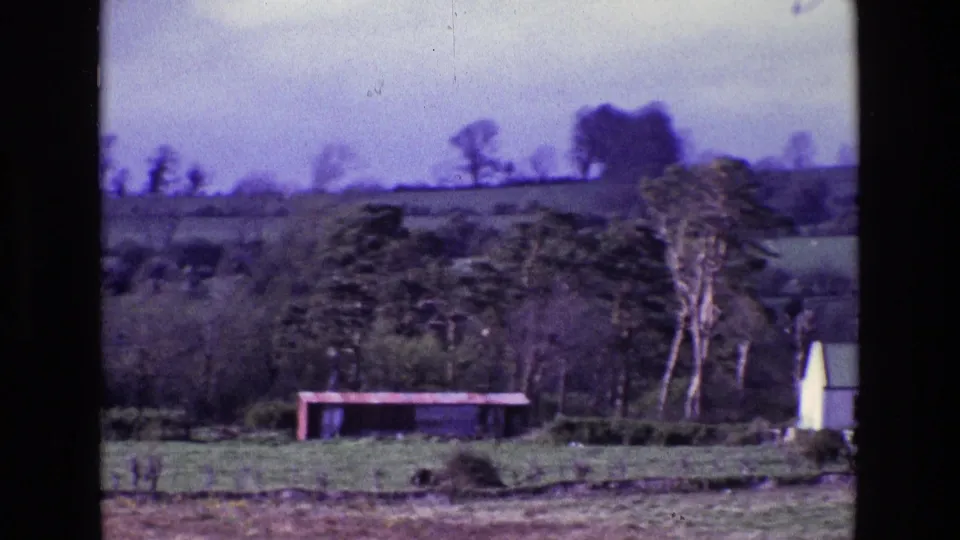 1969 Ireland countryside frame with a green field, trees, a low red structure, and part of a white building at the right edge.