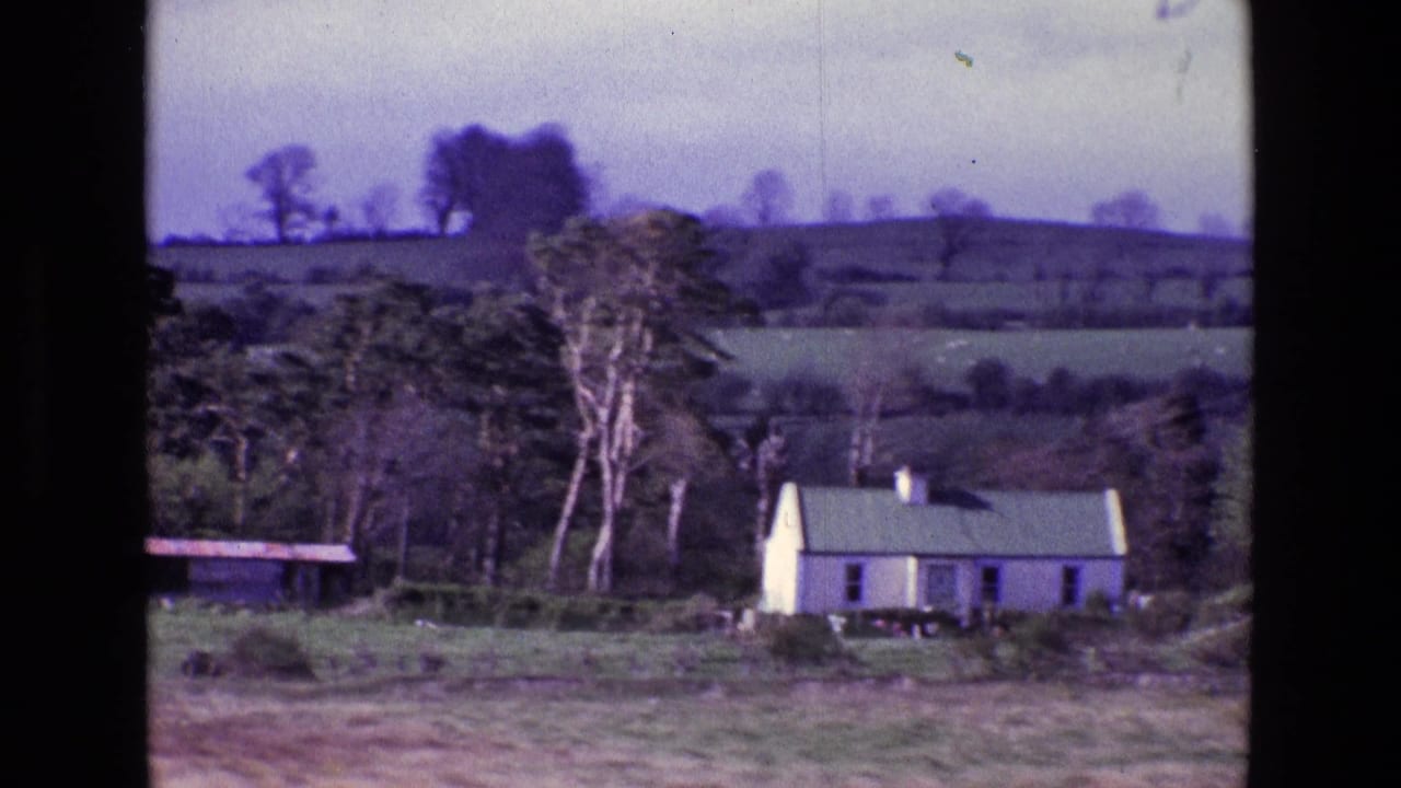 1969 8mm view of a white farmhouse in the Irish countryside, with hedged fields, bare trees, and low hills behind it.