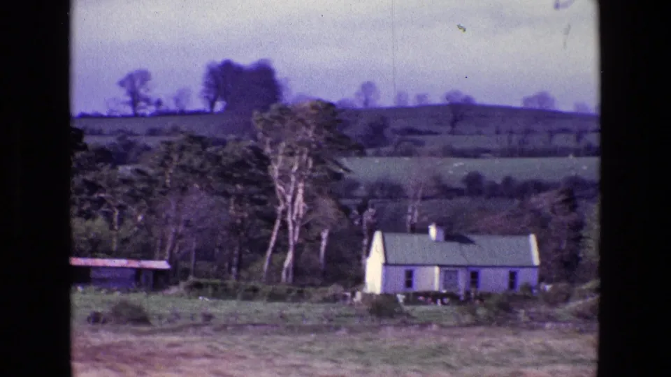 1969 8mm view of a white farmhouse in the Irish countryside, with hedged fields, bare trees, and low hills behind it.