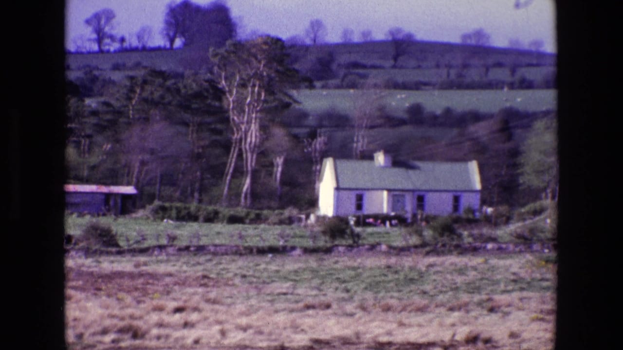 A white rural farmhouse appears in a 1969 Ireland home-movie frame, set below trees, hedgerows, and layered green hills.