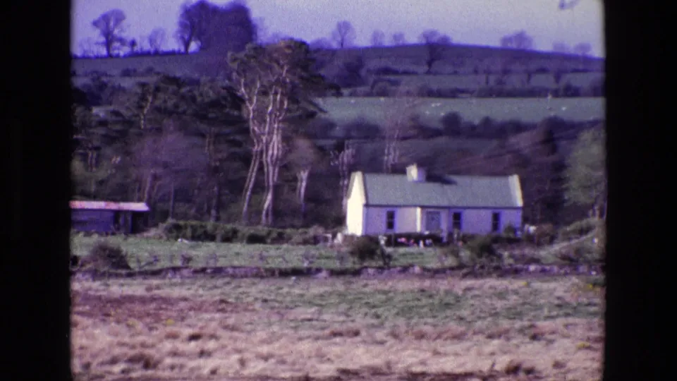 A white rural farmhouse appears in a 1969 Ireland home-movie frame, set below trees, hedgerows, and layered green hills.