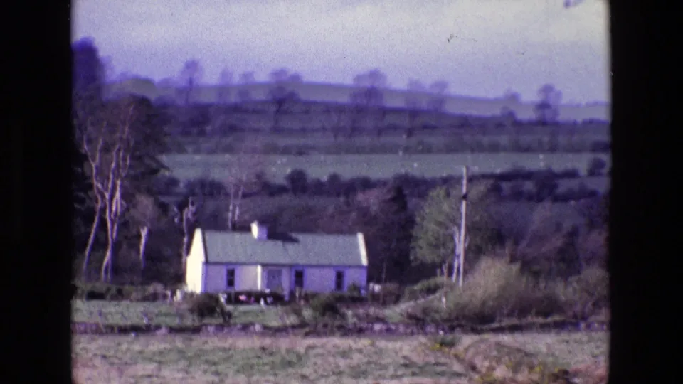 Handheld 8mm frame of a white farmhouse in rural Ireland, with rough foreground grass, hedges, trees, and distant hills.
