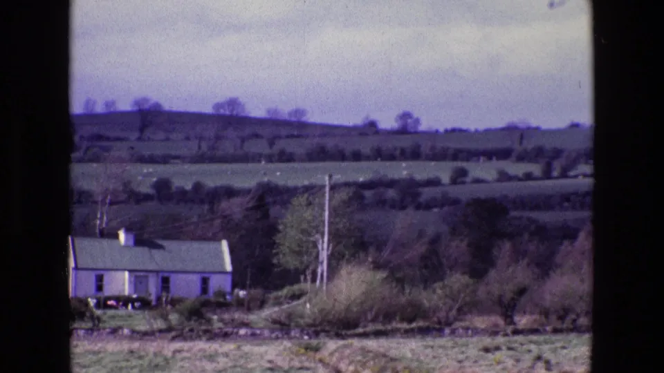 End frame from the 1969 Irish countryside shot, showing a white farmhouse beneath broad fields, hedgerows, bare trees, and cloudy sky.